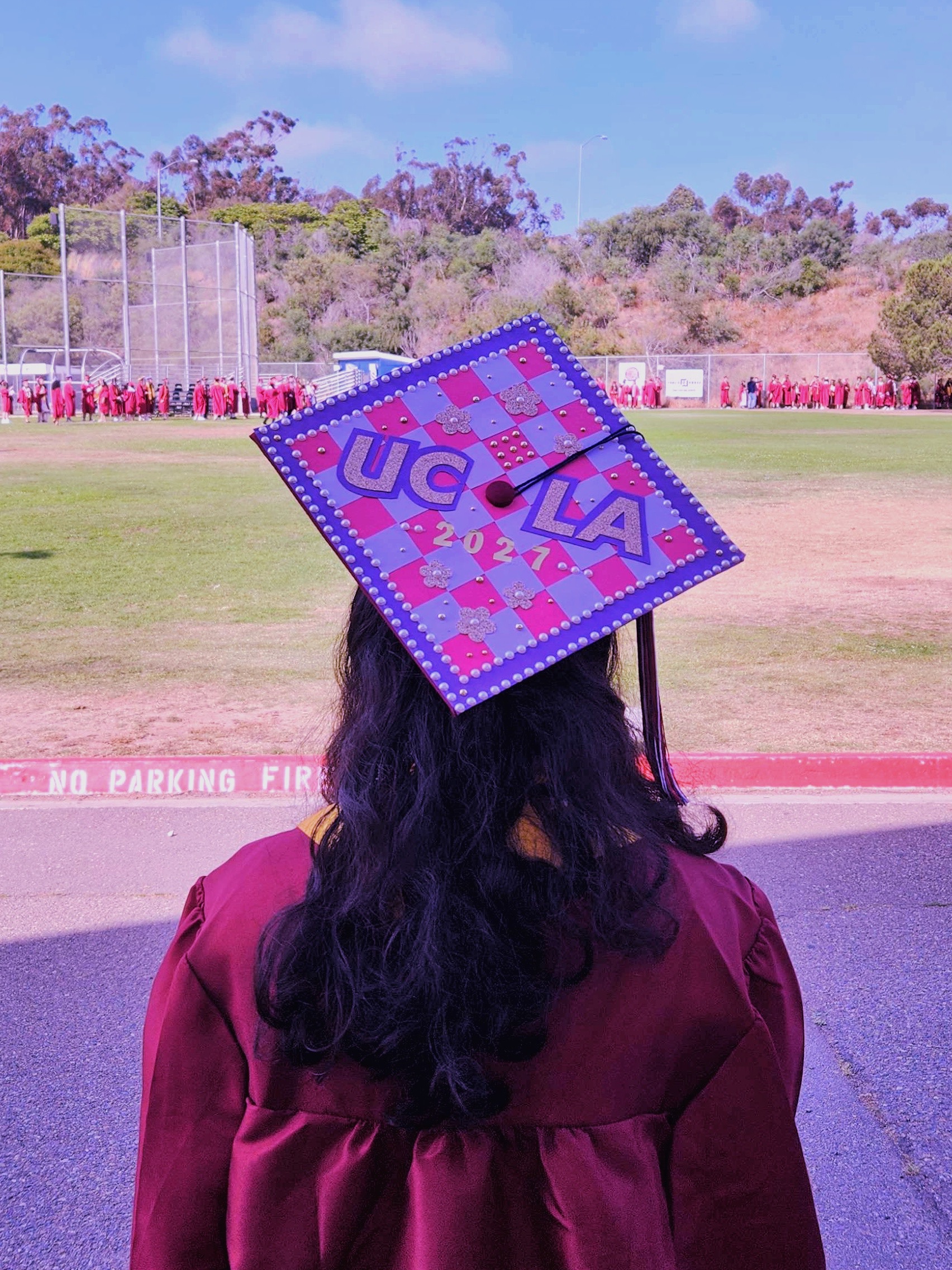 A graduation picture with the back of my graduation cap in the foreground. The design says 'UCLA 2027' and is made up of cut gold, dark purple, light purple, and bright pink cardstock.